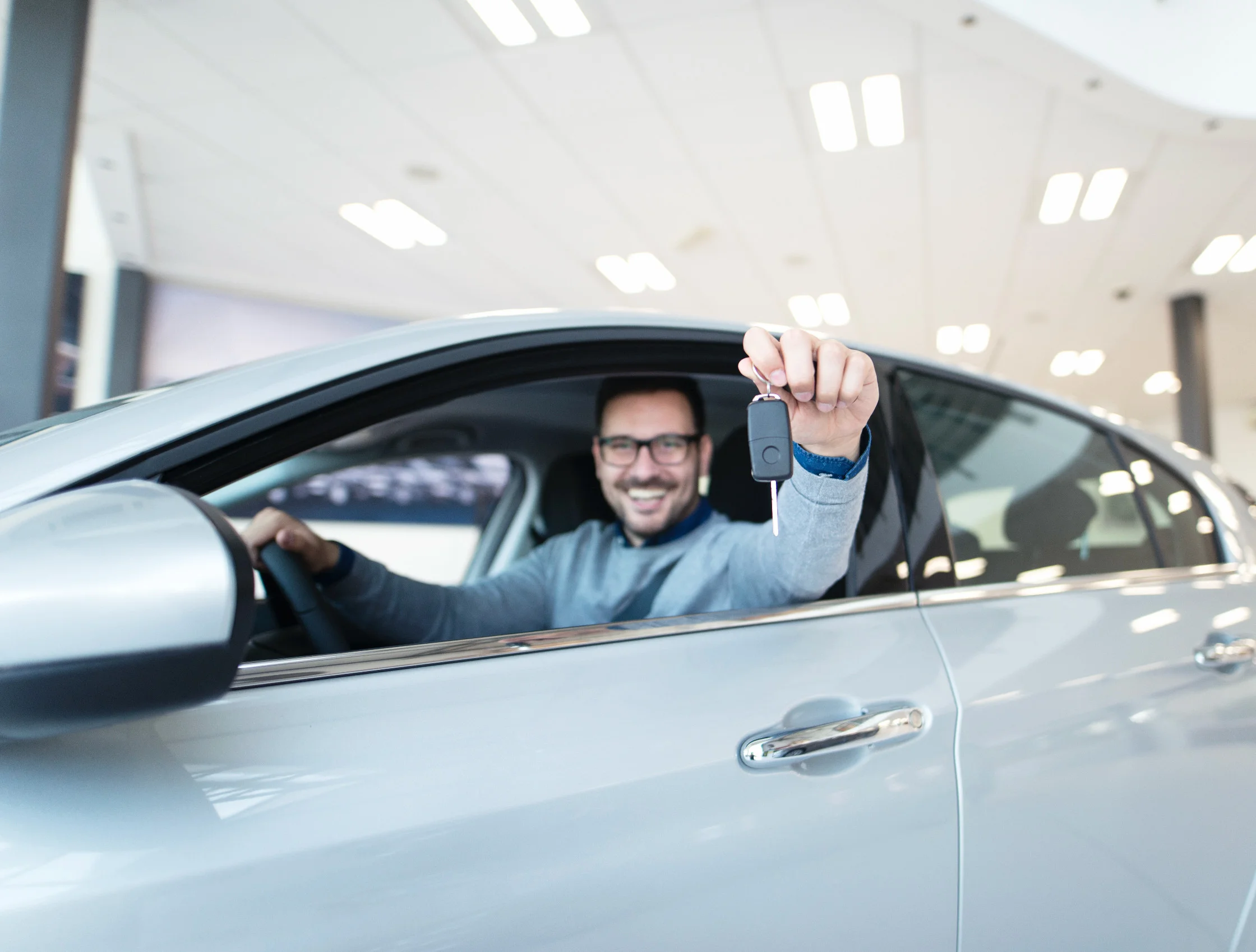 Hombre sonriente con lentes sentado al volante de auto gris sosteniendo las llaves del vehículo