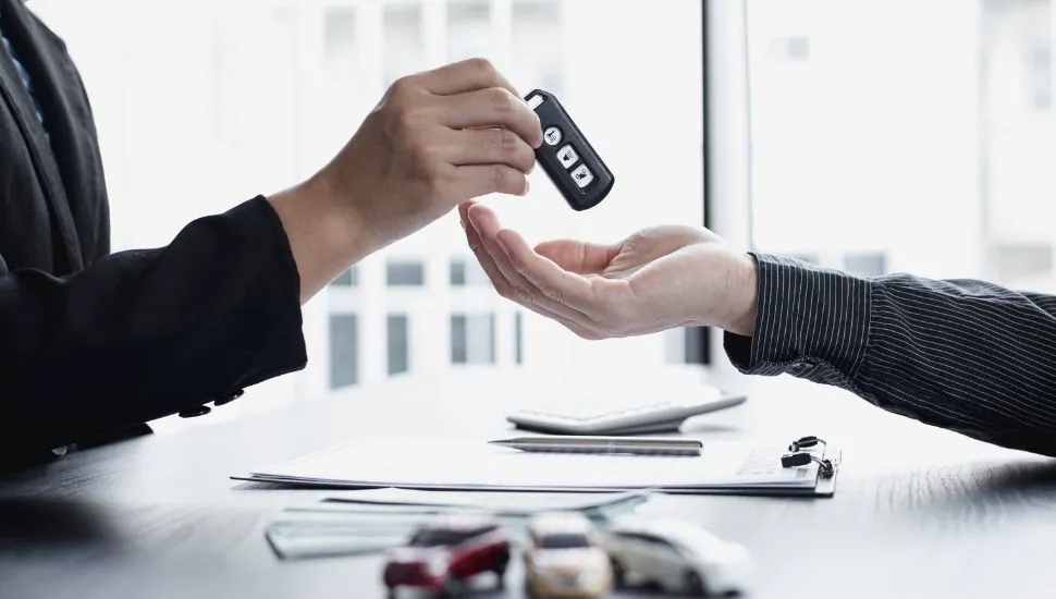 Manos entregando llaves de vehículo sobre mesa con documentos y miniatura de auto amarillo