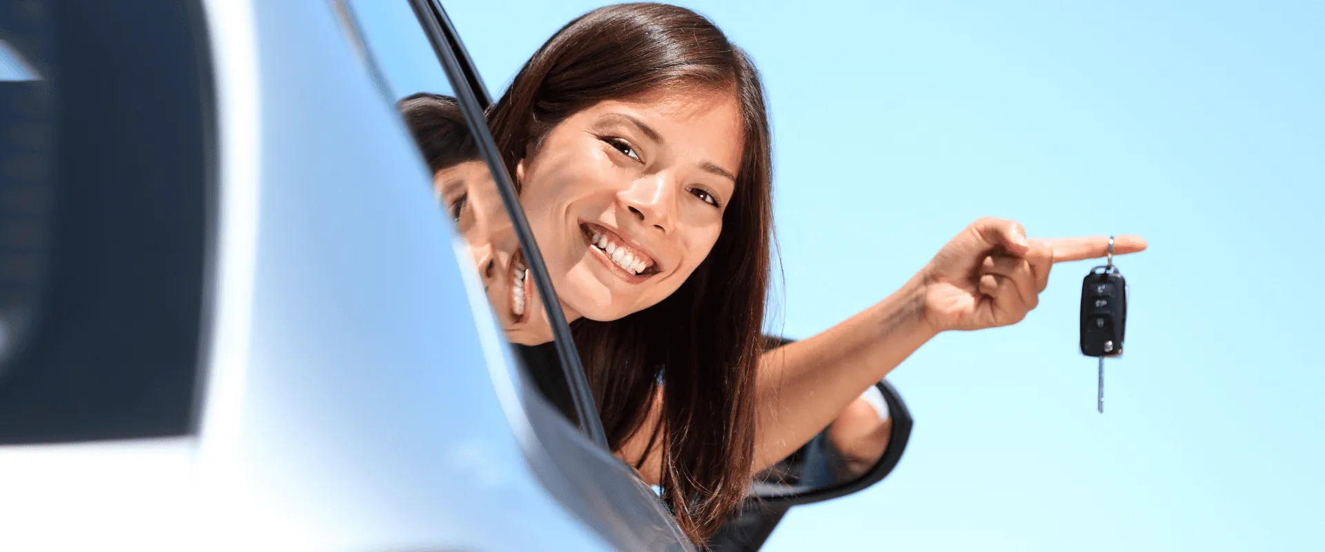 Mujer sonriente asomada por la ventana de un auto sosteniendo las llaves del carro con fondo de cielo azul