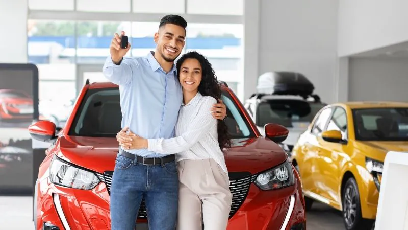 Pareja sonriente en concesionario mostrando las llaves de su auto rojo recién comprado