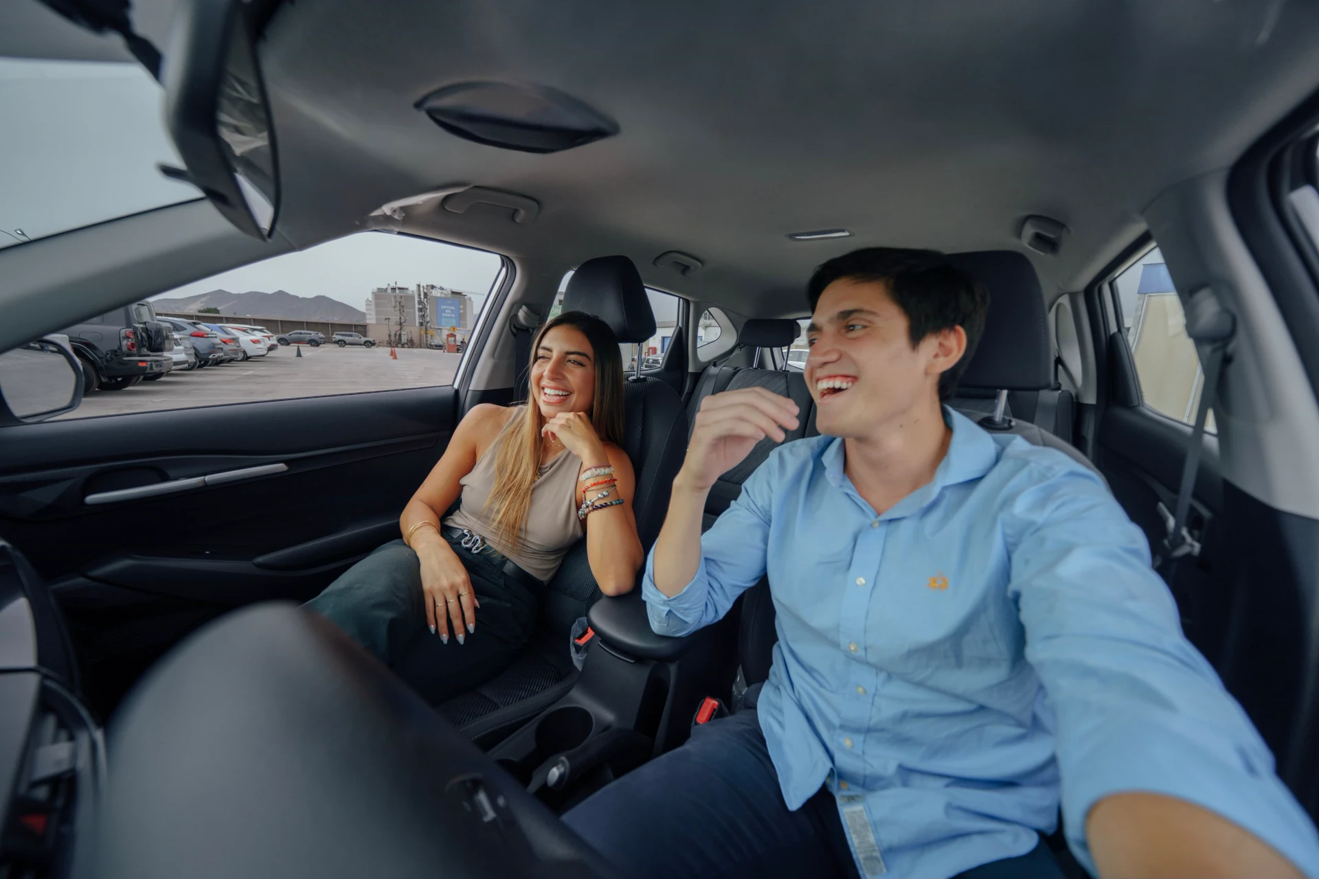 Pareja joven sonriendo sentada en los asientos traseros de un auto seminuevo con estacionamiento de fondo